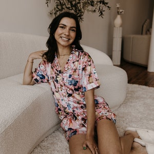 May include: A woman wearing a pink and white pyjama set with a dog print. The pyjamas have short sleeves and shorts. She is sitting on a white fluffy rug in front of a white sofa.