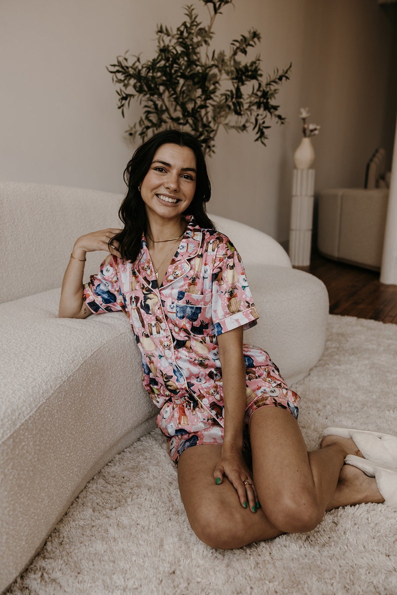 May include: A woman wearing a pink and white pyjama set with a dog print. The pyjamas have short sleeves and shorts. She is sitting on a white fluffy rug in front of a white sofa.