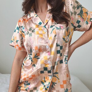 May include: A peach-coloured satin pyjama set with a checkered pattern and a citrus fruit print. The set features a short-sleeved top with a button-up front and matching shorts.