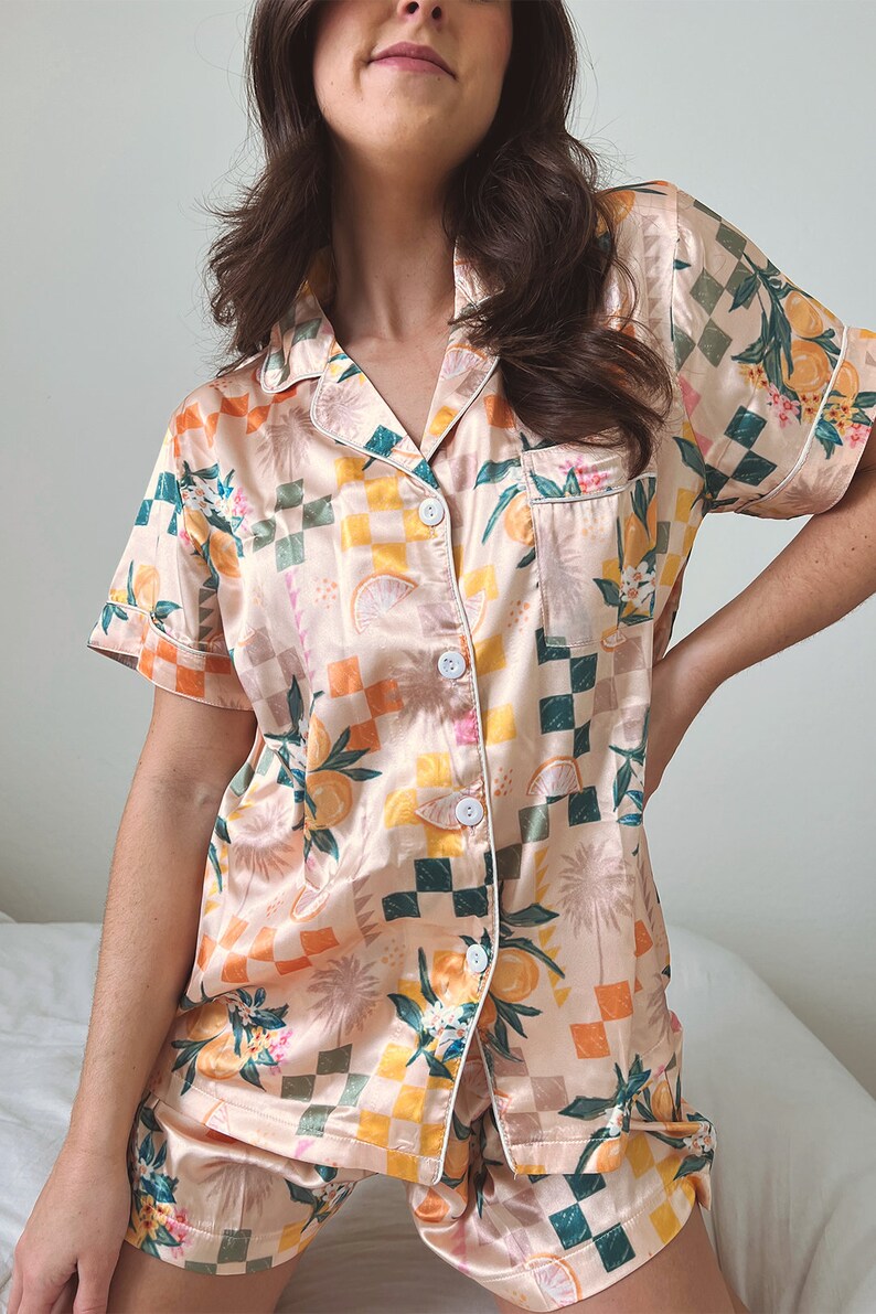 May include: A peach-coloured satin pyjama set with a checkered pattern and a citrus fruit print. The set features a short-sleeved top with a button-up front and matching shorts.