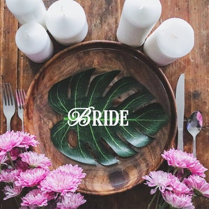 May include: A wooden plate with a green leaf and the word "BRIDE" in white letters. The plate is surrounded by white candles and pink flowers.