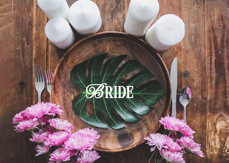 May include: A wooden plate with a green leaf and the word "BRIDE" in white letters. The plate is surrounded by white candles and pink flowers.