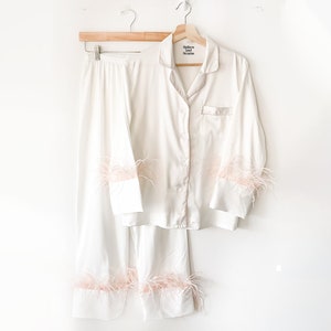 May include: White satin pyjama set with pink feather trim on the cuffs and hem. The top has a button-up front and a small label that reads "Sleeper and The Party".