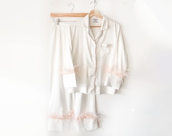 May include: White satin pajama set with pink feather trim on the cuffs and hem. The top has a button-up front and a small label that reads "Sleeper and The Party".