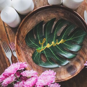 May include: A wooden plate with a green leaf with the name "Laurie" written in gold letters. The plate is surrounded by white candles and pink flowers.