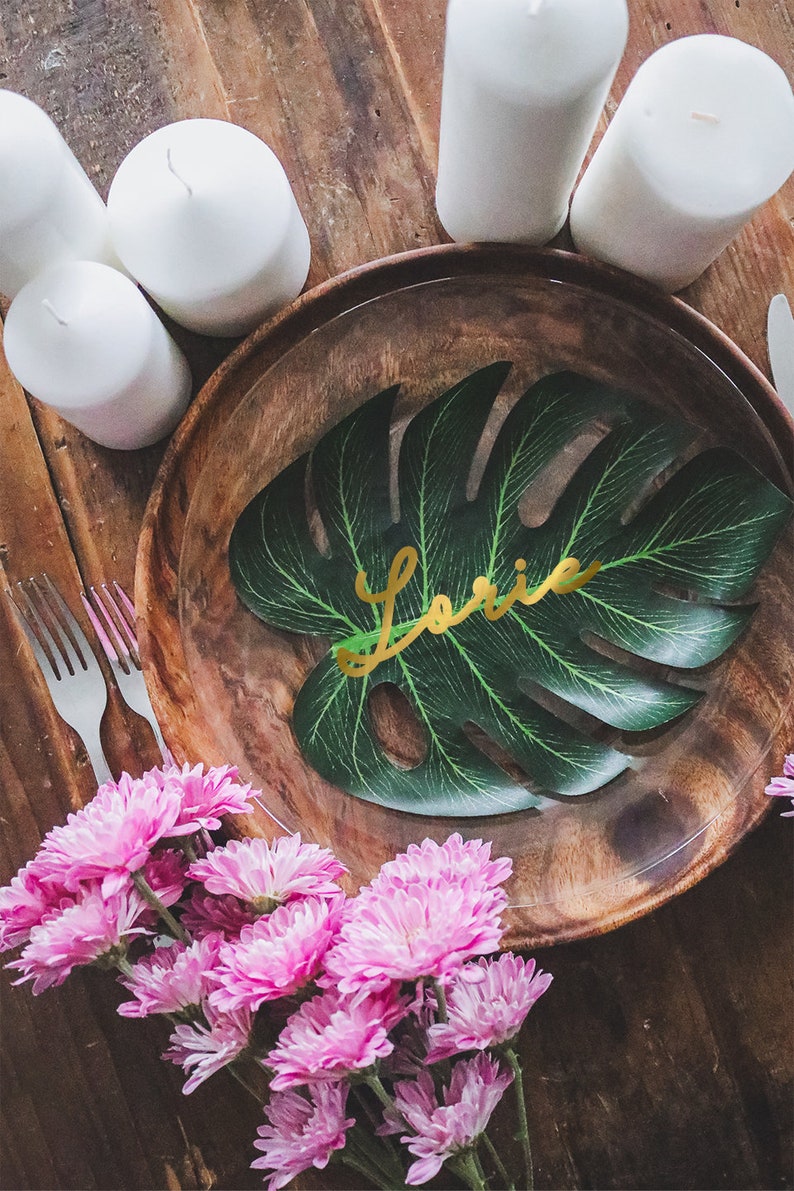 May include: A wooden plate with a green leaf with the name "Laurie" written in gold letters. The plate is surrounded by white candles and pink flowers.