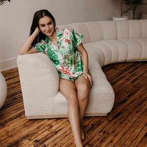 May include: A two-piece pajama set featuring a short-sleeved button-down shirt and matching shorts. The fabric is white with a vibrant tropical print consisting of large green monstera leaves and bright pink hibiscus flowers.
