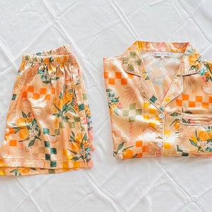 May include: A peach-coloured pyjama set with a checkered and floral pattern featuring oranges and leaves. The top has a collared neckline and a button closure. The shorts have an elastic waistband.