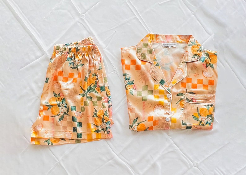 May include: A peach-coloured pyjama set with a checkered and floral pattern featuring oranges and leaves. The top has a collared neckline and a button closure. The shorts have an elastic waistband.
