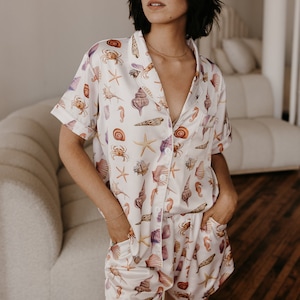 Puede incluir: Conjunto de pijama de satén blanco con una parte superior de manga corta y pantalones cortos. La tela presenta un estampado colorido de conchas marinas, estrellas de mar y caballitos de mar.