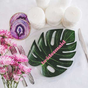 May include: A tropical-themed table setting with a green monstera leaf place card that says "bridesmaid" in pink letters. The table is set with white candles and agate coasters.
