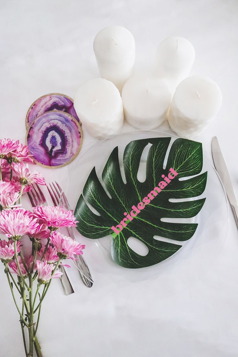 May include: A tropical-themed table setting with a green monstera leaf place card that says "bridesmaid" in pink letters. The table is set with white candles and agate coasters.