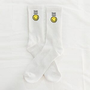 Puede incluir: Un par de calcetines blancos con una carita sonriente amarilla y el texto "best man" en cada calcet&iacute;n.