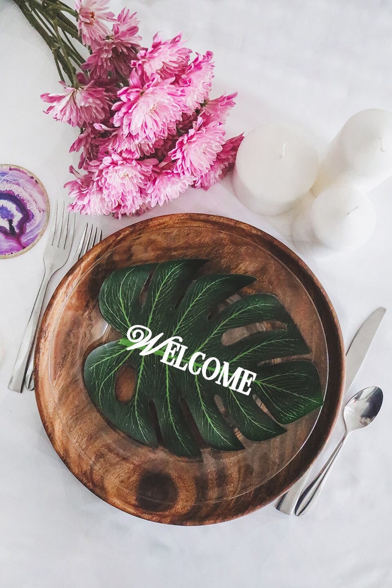 May include: A wooden plate with a green leaf with the word "WELCOME" on it. The plate is set on a white tablecloth with pink flowers and white candles.