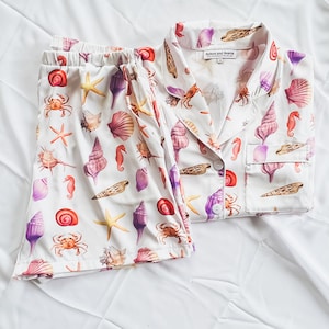 May include: White pajamas with a seashell print. The top has a button-up collar and a pocket on the left side. The bottom is a pair of shorts with an elastic waistband.