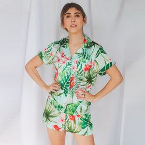 May include: A two-piece pajama set featuring a short-sleeved button-down shirt and matching shorts. The fabric is a light mint green color decorated with a vibrant tropical print of large dark green monstera leaves and bright pink flowers.