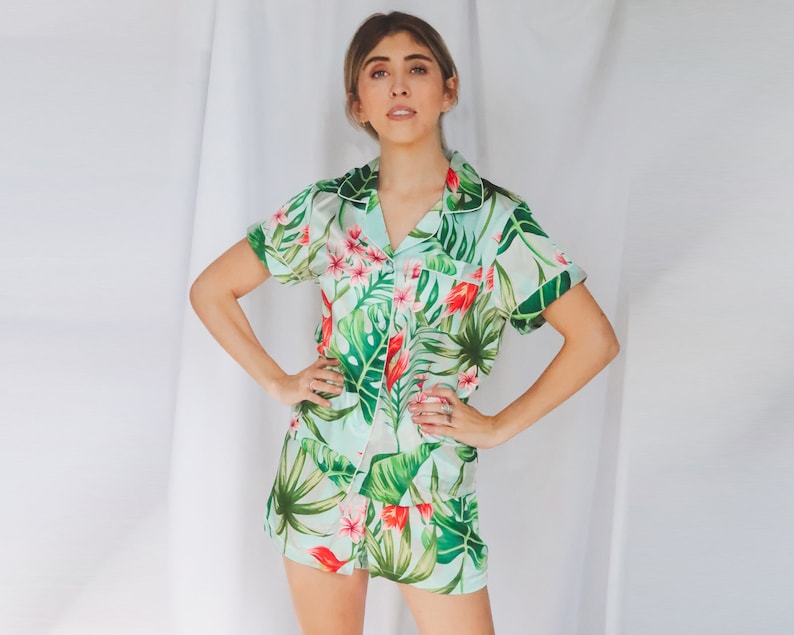 May include: A two-piece pajama set featuring a short-sleeved button-down shirt and matching shorts. The fabric is a light mint green color decorated with a vibrant tropical print of large dark green monstera leaves and bright pink flowers.