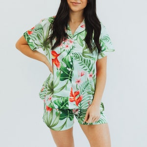 May include: A two-piece pajama set featuring a short-sleeved button-down shirt and matching shorts. The set is light mint green with a vibrant tropical print of large green monstera leaves, palm fronds, and bright red hibiscus flowers.