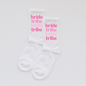 May include: Two white socks with the words "bride tribe" printed on them in pink. The words are stacked on top of each other, with "bride" in a larger font than "tribe".