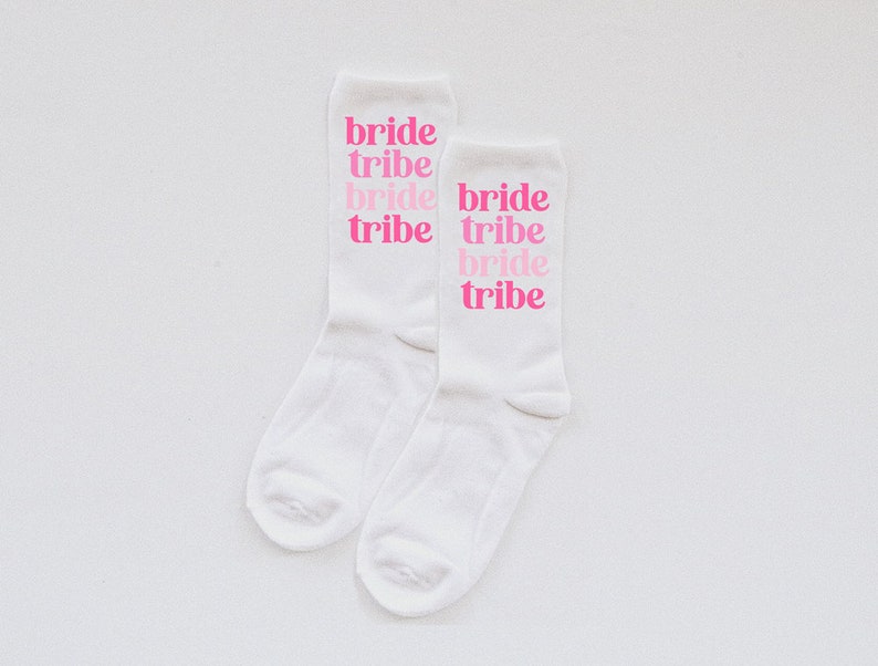 May include: Two white socks with the words "bride tribe" printed on them in pink. The words are stacked on top of each other, with "bride" in a larger font than "tribe".