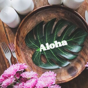 May include: A wooden plate with a green tropical leaf with the word "Aloha" on it. Pink flowers are in the foreground.