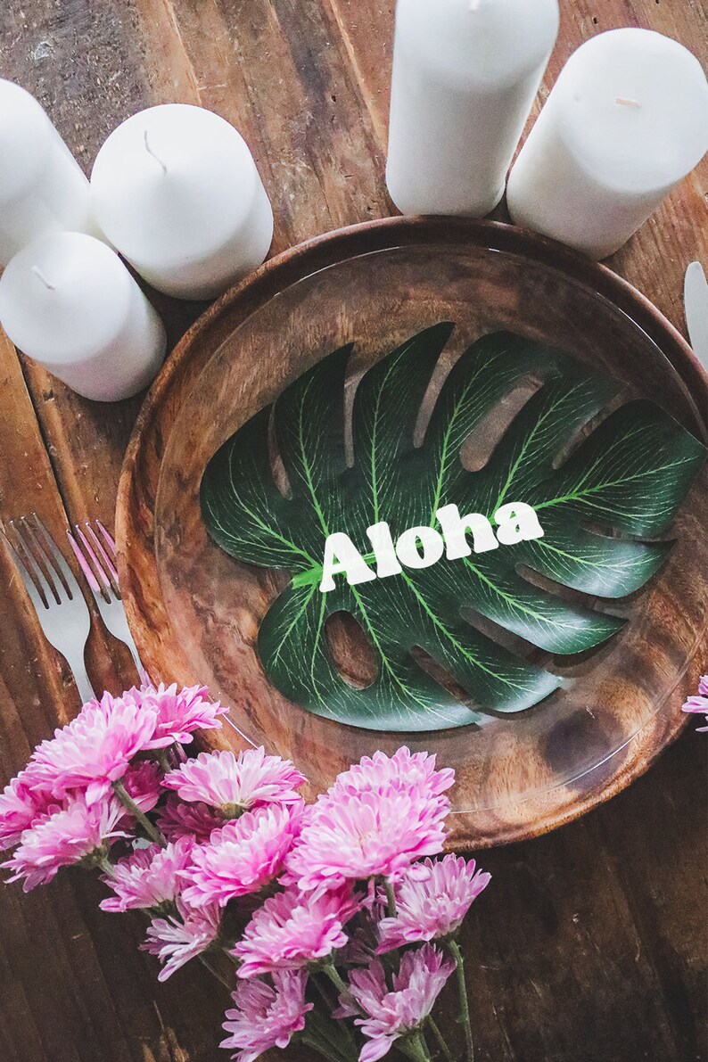 May include: A wooden plate with a green tropical leaf with the word "Aloha" on it. Pink flowers are in the foreground.