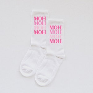 May include: A pair of white socks with the word "MOH" repeated four times in pink lettering. The lettering is in a gradient of pink, with the top "MOH" being the lightest pink and the bottom "MOH" being the darkest pink.