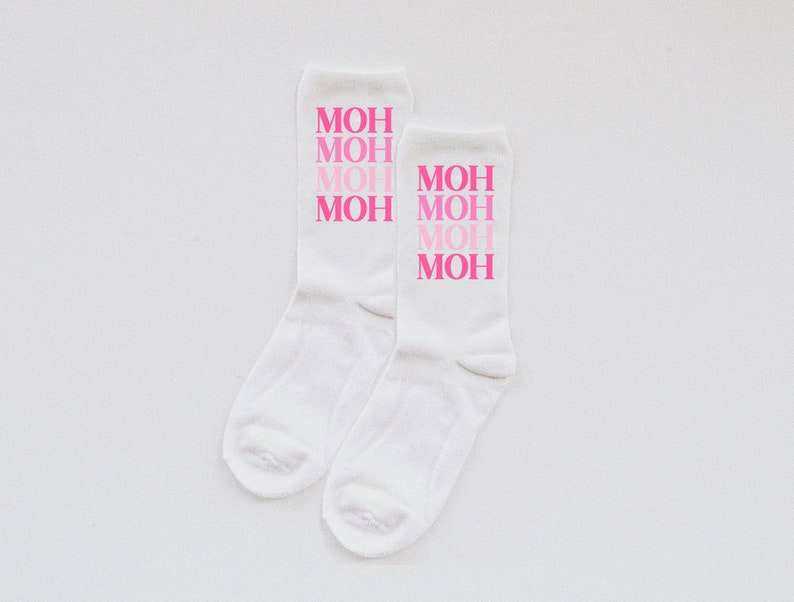 May include: A pair of white socks with the word "MOH" repeated four times in pink lettering. The lettering is in a gradient of pink, with the top "MOH" being the lightest pink and the bottom "MOH" being the darkest pink.