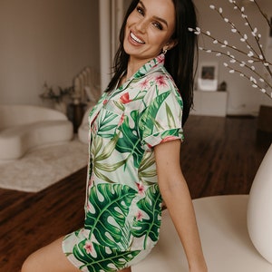 May include: A two-piece pajama set featuring a short-sleeved button-down shirt and matching shorts. The fabric is light-colored with a vibrant green tropical leaf pattern and small pink floral accents throughout the garment.
