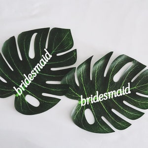 May include: Two green monstera leaves with the word "bridesmaid" written on each leaf in white.