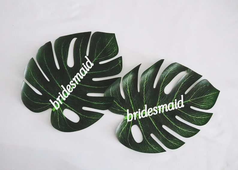 May include: Two green monstera leaves with the word "bridesmaid" written on each leaf in white.