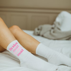 May include: White socks with pink text that reads "Brides maid Brides maid". The socks are on a white bed.