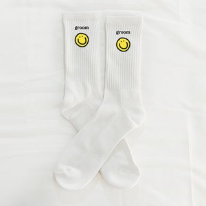 Puede incluir: Un par de calcetines blancos con una carita sonriente amarilla y la palabra "groom" impresa en cada calcet&iacute;n.