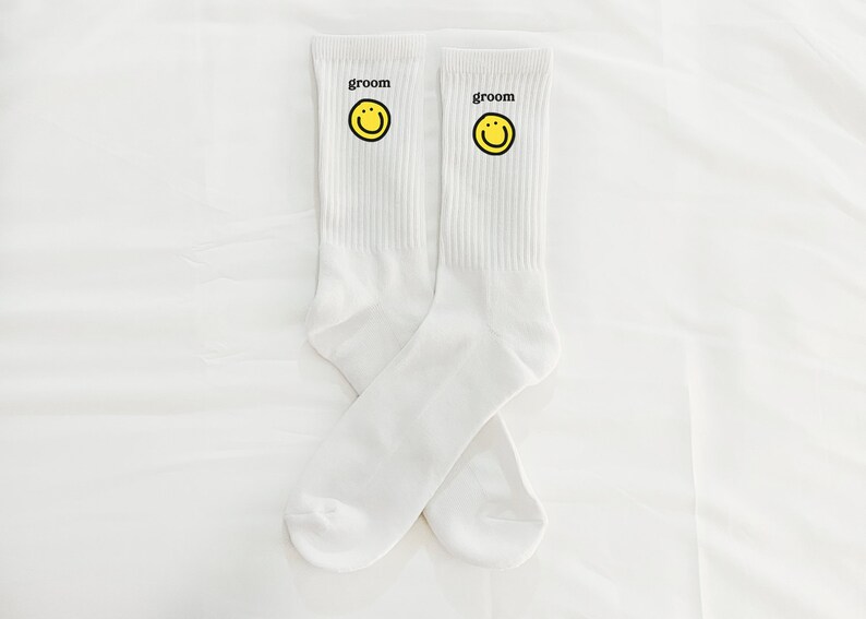 Puede incluir: Un par de calcetines blancos con una carita sonriente amarilla y la palabra "groom" impresa en cada calcet&iacute;n.