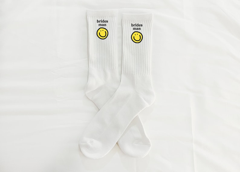 Puede incluir: Un par de calcetines blancos con una carita sonriente amarilla y el texto "brides man" en cada calcet&iacute;n.