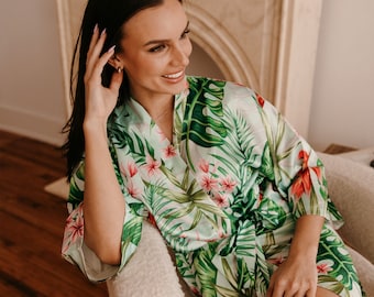 Puede incluir: Una mujer lleva una bata de satén verde y blanca con estampado floral tropical. La bata tiene un cinturón de lazo y está hecha de una tela suave y sedosa.