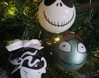 May include: Three Christmas tree ornaments featuring characters from The Nightmare Before Christmas. The ornaments are round, with Jack Skellington, Sally, and a third character designs. The ornaments are hanging on a green Christmas tree.