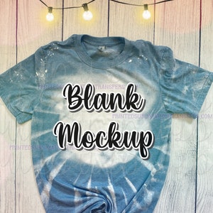 May include: A blue tie-dye t-shirt with the words "Blank Mockup" in black script. The shirt has a bleached effect and is displayed on a light wood background. The shirt is a crew neck and has short sleeves.