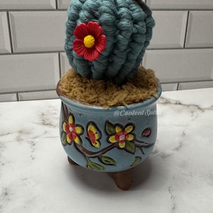 May include: A blue crocheted cactus with red flowers in a blue and white ceramic pot with floral designs. The pot has three brown feet.