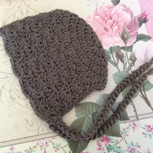 Puede incluir: Un gorro de bebé de ganchillo marrón con lazos largos. El gorro está hecho con un patrón de punto sencillo y tiene una sensación suave y acogedora.