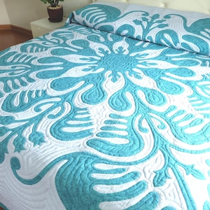 Op de afbeelding: Een bed met een wit en turquoise sprei. De sprei heeft een groot bloemmotief in turquoise, met gedetailleerde quiltsteken. Het bed is opgemaakt en klaar voor gebruik.
