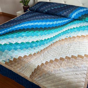 Può includere: Un letto coperto da una trapunta con un design a tema spiaggia. La trapunta presenta una sfumatura di colori, tra cui tonalità di blu, verde e beige, che ricordano l'oceano e la sabbia. La trapunta è realizzata in tessuto con un motivo geometrico.