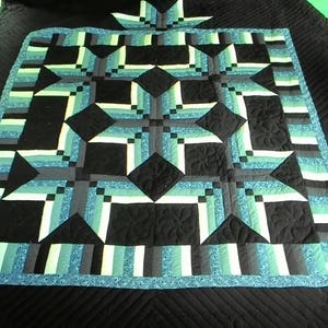 May include: A handmade quilt with a geometric star pattern in shades of black, teal, gray, and white. The quilt features a central star design surrounded by a border of alternating stripes. The quilt is made of fabric and has a quilted texture.