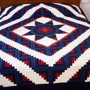 May include: A handcrafted quilt featuring a central star design in navy blue, red, and white. The quilt's pattern includes a diamond shape with a star in the center, surrounded by a zig-zag pattern. The quilt is on a bed.