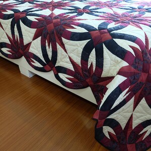 May include: A detailed quilt with a repeating pattern of interlocking rings and starbursts in burgundy, black, and cream. The quilt is draped over a bed, showcasing its intricate design and craftsmanship. The quilt's edge is scalloped.