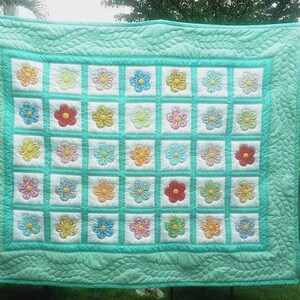 Peut inclure: Une courtepointe faite &agrave; la main avec un motif floral. La courtepointe pr&eacute;sente une grille de carr&eacute;s blancs, chacun contenant une fleur color&eacute;e. Les fleurs sont dans les tons de rouge, jaune, bleu et orange. La courtepointe a une bordure et un dos turquoise.
