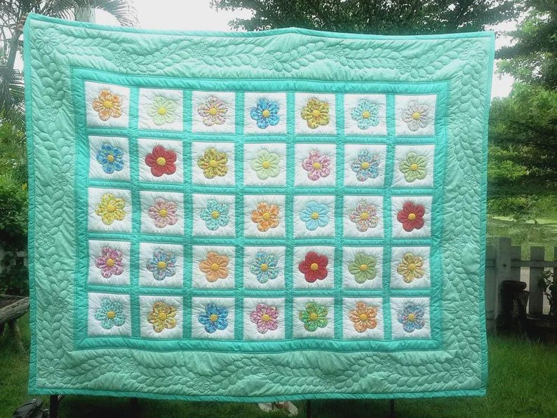 Peut inclure: Une courtepointe faite &agrave; la main avec un motif floral. La courtepointe pr&eacute;sente une grille de carr&eacute;s blancs, chacun contenant une fleur color&eacute;e. Les fleurs sont dans les tons de rouge, jaune, bleu et orange. La courtepointe a une bordure et un dos turquoise.