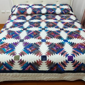 May include: A colourful, handmade quilt featuring a repeating geometric pattern in shades of blue, purple, red, and green on a white background. The quilt is on a bed with a white headboard and matching pillow.