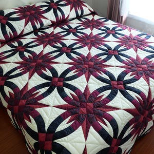 May include: A handmade quilt with a striking pattern. The quilt features a repeating design of interlocking circles and starbursts in burgundy, navy, and cream. The quilt is placed on a bed with a white headboard and a wooden floor.