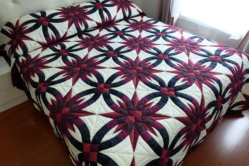 May include: A handmade quilt with a striking pattern. The quilt features a repeating design of interlocking circles and starbursts in burgundy, navy, and cream. The quilt is placed on a bed with a white headboard and a wooden floor.